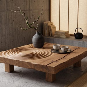Op de afbeelding: Een lage, vierkante houten salontafel met een uitgesneden rimpelpatroon op het oppervlak. De tafel heeft vier blokpoten en een natuurlijke houtnerfafwerking. Een donkergrijze vaas met een kale tak staat naast een dienblad met twee kopjes.