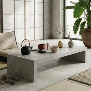 Puede incluir: Una mesa de centro rectangular baja de hormigón con una silla de suelo negra con cojín beige. Sobre la mesa hay una tetera negra con asa de bambú, un cuenco rojo, un cuenco beige pequeño, un batidor de madera, un bote dorado y un jarrón pequeño.