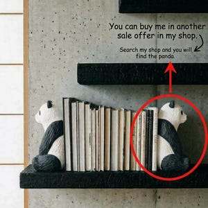 May include: Two black and white panda-shaped bookends positioned on a black shelf, holding a row of books between them. The panda figures are depicted in a seated posture, facing inward toward the books, with detailed black and white coloring.