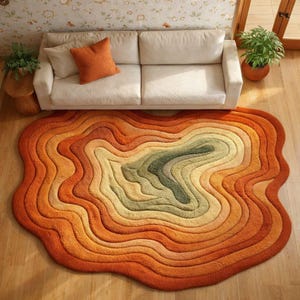Könnte beinhalten: Ein einzigartiger, geschichteter Teppich in Orange-, Gelb- und Grüntönen, der einer topografischen Karte ähnelt. Der Teppich liegt auf einem Holzboden, mit einem weißen Sofa und einem orangefarbenen Kissen im Hintergrund. Das Design des Teppichs verleiht dem Raum eine warme, einladende Atmosphäre.