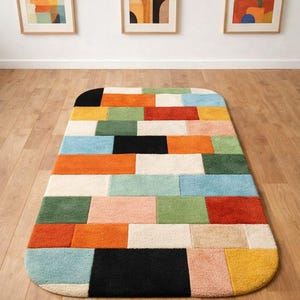 Könnte beinhalten: Ein rechteckiger Teppich mit abgerundeten Ecken, der ein Patchwork-Design in verschiedenen Farben aufweist. Der Teppich enthält Blöcke in Schwarz, Orange, Grün, Blau und Gelb. Drei gerahmte abstrakte Kunstwerke hängen an der weißen Wand über dem Teppich.