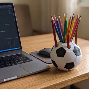 May include: A spherical pencil holder designed to resemble a black and white football, featuring multiple circular openings on the top surface. The holder is filled with an assortment of sharpened coloured pencils in various shades.
