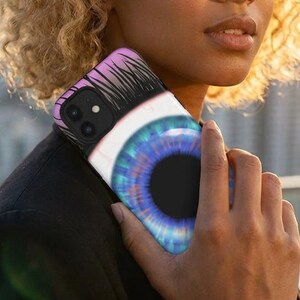 May include: A smartphone case featuring a large, stylized eye design. The eye has a blue iris with a black pupil, surrounded by white and a pink and black eyelash design. The case is held against a blurred cityscape background.