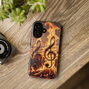 May include: Black phone case featuring a fiery design with two musical treble clef symbols and musical staff lines. The background shows flames and embers, creating a dramatic and artistic look. The case is shown on a wooden surface.