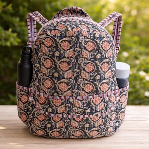 Puede incluir: Mochila acolchada con un estampado floral en azul marino, melocotón y azul. La mochila tiene dos bolsillos laterales, uno con una botella de agua negra y otro con un recipiente blanco y azul. Las correas y el ribete presentan un diseño a rayas.