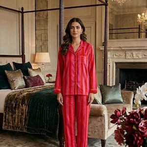 May include: A woman wearing a red and pink striped pajama set. The long-sleeved top and pants have white trim. The bedroom features a four-poster bed, a sofa, and a fireplace. Chandeliers hang from the ceiling.
