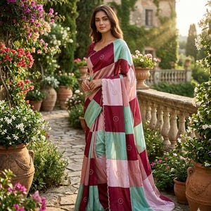 May include: A patchwork saree in shades of burgundy, pink, and mint green. The saree features a geometric pattern with circular accents. The woman is standing in a garden setting with flowers and terracotta pots.