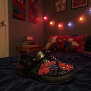 May include: A black clog shoe featuring a pivoting heel strap adorned with silver-toned metal studs. The upper surface is embellished with black rhinestones and two distinct Spider-Man character patches in red and blue colors.