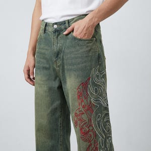May include: Faded green denim jeans with a wide leg cut. The jeans feature a large embroidered design in red and white on the lower leg. The jeans have a classic five-pocket design and a button and zipper closure.