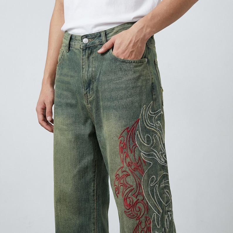 May include: Faded green denim jeans with a wide leg cut. The jeans feature a large embroidered design in red and white on the lower leg. The jeans have a classic five-pocket design and a button and zipper closure.