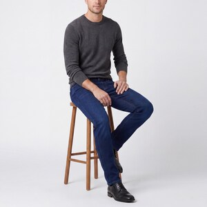 May include: A person wearing a dark gray crew neck sweater and dark blue jeans, seated on a wooden stool. The person is also wearing black leather dress shoes. The background is plain white.