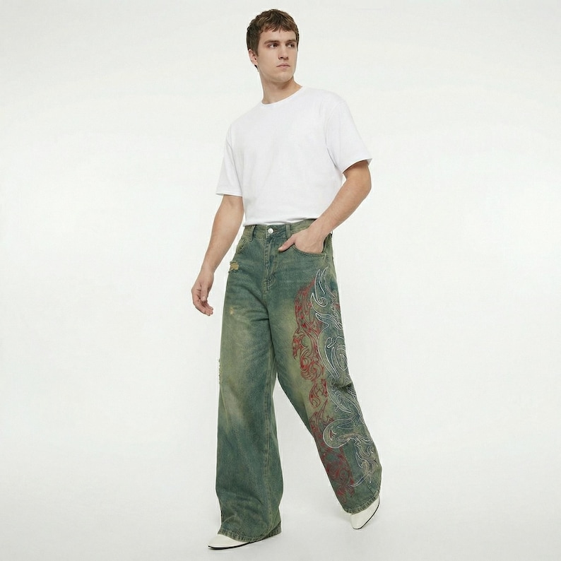 May include: Wide-leg, distressed denim jeans with a faded green wash and a large, ornate red and blue design on the right leg. The jeans have a classic five-pocket design and a button and zipper closure.