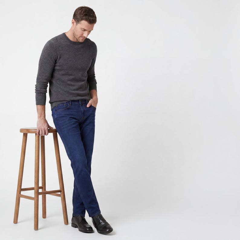 May include: A charcoal gray crew neck sweater and dark blue jeans. The person is wearing black leather boots and leaning on a wooden stool. The background is plain white.