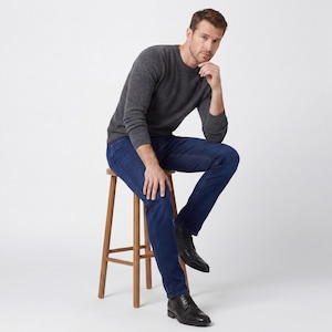 May include: A man wearing a charcoal gray crew neck sweater and dark blue jeans, seated on a wooden stool. He is also wearing black leather boots. The background is plain white.