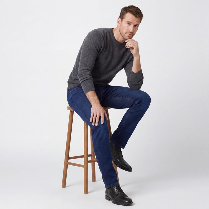 May include: A man wearing a charcoal gray crew neck sweater and dark blue jeans, seated on a wooden stool. He is also wearing black leather boots. The background is plain white.