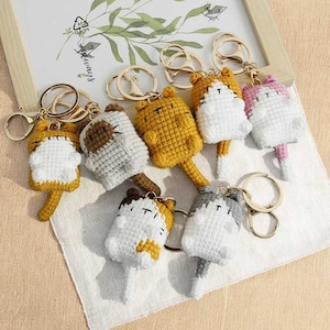 Może przedstawiać: Kolekcja ręcznie robionych breloków do kluczy w kształcie kota wykonanych na szydełku w różnych kolorach, w tym żółtym, białym, szarym i różowym. Każdy brelok ma wzór kota z ogonem i złotym kółkiem.