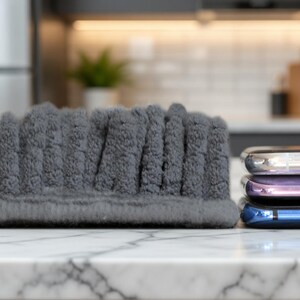 May include: A grey, textured headband with a ribbed design sits on a marble surface. The headband is next to a stack of three smartphones with silver, purple, and blue backs. The background is a blurred kitchen.