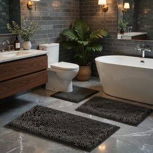 May include: A contemporary bathroom with a white bathtub, toilet, and vanity. Three dark grey rectangular bath mats are on the floor. The room features grey brick-style tiles, a large plant, and gold-toned wall lights.