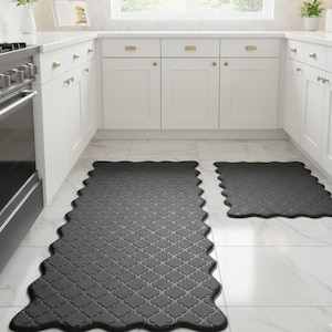 May include: Two dark gray kitchen mats with a scalloped edge and a repeating quatrefoil pattern. One is a long runner, and the other is a smaller square mat. The mats are on a white tile floor in a kitchen.