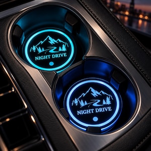 May include: Two circular car cup holder inserts featuring a bright blue LED glow. Each insert displays a graphic of a mountain range, pine trees, and a winding road with the text NIGHT DRIVE printed in white below the landscape design.