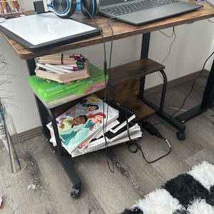 May include: A mobile desk with a laptop, headphones, and a whiteboard on top. The desk has a lower shelf with art supplies and books. The desk is brown with black metal legs and wheels. A black and white checkered rug is on the floor.