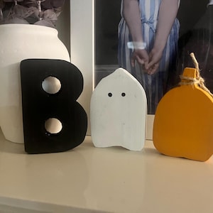 May include: A set of three decorative wooden cutouts consisting of a black letter B, a white ghost shape with two black circular eyes, and an orange pumpkin shape with a brown twine bow tied around the stem, arranged in a horizontal row.
