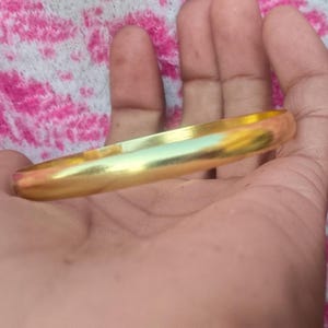 May include: A gold-colored bangle bracelet with a smooth, polished surface. The bracelet is a solid band, reflecting light with a warm, golden hue. The jewelry is a simple, elegant design.