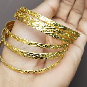 Może przedstawiać: Kolekcja złotych bransoletek ze skomplikowanymi wzorami kwiatowymi. Bransoletki są ułożone jedna na drugiej, prezentując ich szczegółowy design. Biżuteria jest prezentowana na neutralnym tle.