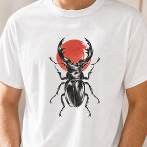 Può includere: Una t-shirt bianca a maniche corte con un'illustrazione grafica di un cervo volante nero centrata sul petto. Dietro il coleottero si trova una forma circolare rossa piena. Il design è realizzato con uno stile artistico dettagliato a inchiostro.