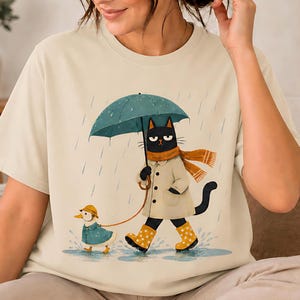 May include: A cream-colored t-shirt featuring a graphic of a black cat wearing a beige trench coat, a mustard yellow scarf, and yellow polka-dot rain boots. The cat holds a teal umbrella while walking a small duckling in a blue raincoat.
