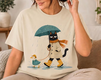 Peut inclure: Un t-shirt couleur crème avec une illustration d'un chat noir portant un trench-coat beige, une écharpe jaune moutarde et des bottes de pluie jaunes à pois. Le chat tient un parapluie bleu sarcelle et promène un caneton en imperméable bleu.