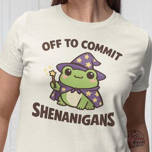 May include: Cream t-shirt with a cartoon frog wizard design. The frog is green, wearing a purple hat and cape with gold stars, and holding a wand. Text reads "OFF TO COMMIT SHENANIGANS" in brown.