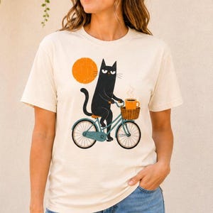 A woman wearing a cream t-shirt with an illustration of a black cat riding a bicycle with a coffee cup in the front basket.