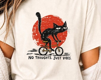 Peut inclure: Un t-shirt couleur crème arborant une illustration d'un chat noir sur un vélo jaune devant un grand cercle rouge. Sous le dessin, le texte "NO THOUGHTS. JUST VIBES." est inscrit dans une police noire, grasse et sans empattement.