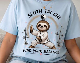 Peut inclure: Un t-shirt bleu clair avec une illustration d'un paresseux brun portant un uniforme d'arts martiaux blanc en position de tai-chi. Au-dessus du paresseux, le texte "SLOTH TAI CHI" et un symbole yin-yang. En dessous, "FIND YOUR BALANCE" et des bambous.