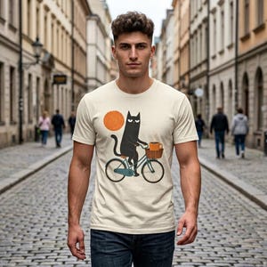 A man walking down a city street wearing a cream t-shirt featuring a black cat riding a bicycle with a cup of coffee.