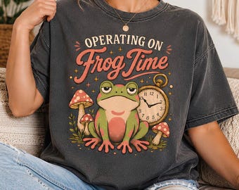 Peut inclure: Un t-shirt gris anthracite avec une impression graphique représentant une grenouille verte assise à côté d'une montre de poche dorée et de deux champignons à pois rouges. Le texte au-dessus indique "OPERATING ON Frog Time" en rouge clair.