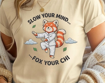 Peut inclure: T-shirt beige avec un panda roux de dessin animé en uniforme d'arts martiaux blanc, pratiquant une pose de Tai Chi. Le texte "SLOW YOUR MIND... ...FOX YOUR CHI" est en noir. Des feuilles vertes et des nuages complètent le design.