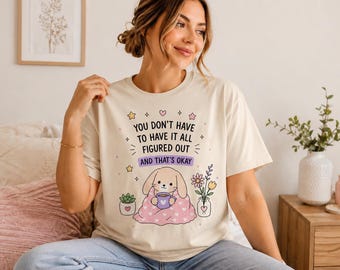 Peut inclure: Un t-shirt couleur crème avec un motif représentant un lapin enveloppé dans une couverture rose à cœurs tenant une tasse. Le texte indique : YOU DON'T HAVE TO HAVE IT ALL FIGURED OUT AND THAT'S OKAY, entouré de petites étoiles, cœurs et fleurs.