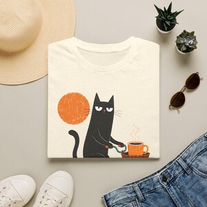 Flat lay of a folded cream t-shirt with a black cat on a bicycle graphic, surrounded by a straw hat, sunglasses, plants, and jeans.