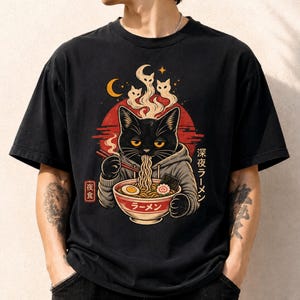 May include: A black t-shirt featuring a graphic of a black cat wearing a gray hoodie, eating ramen from a bowl with chopsticks. Above the cat are three ghost-like cat shapes and a crescent moon. Japanese text reads 深夜ラーメン and ラーメン.