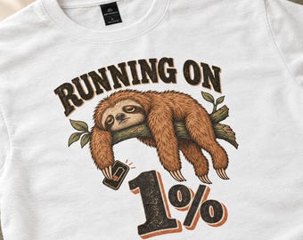 Peut inclure: Un t-shirt blanc avec une illustration d'un paresseux brun suspendu à une branche d'arbre, tenant un smartphone avec une icône de batterie faible. Le texte au-dessus indique RUNNING ON et le texte en dessous indique 1% en lettres noires et orange.