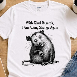 Könnte beinhalten: Weißes T-Shirt mit einer schwarz-weißen Illustration eines Opossums mit einem komischen Gesichtsausdruck. Der Text "With Kind Regards, I Am Acting Strange Again" ist über dem Opossum aufgedruckt. Das Shirt ist von der Marke Gildan.