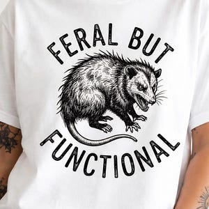 Könnte beinhalten: Weißes T-Shirt mit einer schwarzen Grafik eines Opossums und dem Text "FERAL BUT FUNCTIONAL". Das Opossum ist detailliert dargestellt, mit sichtbarem Fell und Gesichtszügen. Der Text ist fett und ohne Serifen.
