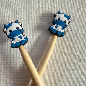 May include: Two pencils topped with blue and white cartoon cow figures. The cow figures have black outlines and are sitting down. The pencils are made of light-colored wood and are crossed over each other.