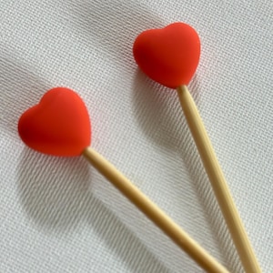 May include: Two red heart-shaped toppers on wooden sticks, set against a white textured background. The toppers are a vibrant red, and the sticks are a natural wood color. The image is a close-up shot.