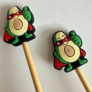 Può includere: Due matite fantasiose con decorazioni a forma di avocado supereroe. Ogni personaggio di avocado indossa un mantello e una maschera rossi, con pelle verde, un nocciolo marrone e un gesto di pollice in su. Le matite hanno fusti in legno naturale.