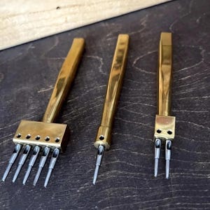 May include: Three brass leather working tools. The tools have a rectangular brass handle and a series of silver-colored prongs. The tools are designed for creating evenly spaced holes in leather for stitching.