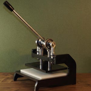 Op de afbeelding: Een zwarte en zilveren handpers met een lange hendel en een witte basis. De pers heeft een zwarte basis en een zilveren hefboomarm met een zwarte handgreep. De pers is ontworpen voor knutselen en andere kleine projecten.
