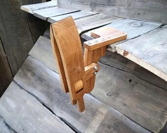 May include: A wooden woodworking vise, constructed from light brown wood, is mounted on a wooden surface. The vise features a curved clamping mechanism and a rectangular base, secured with metal hardware. The background is a weathered wooden structure.